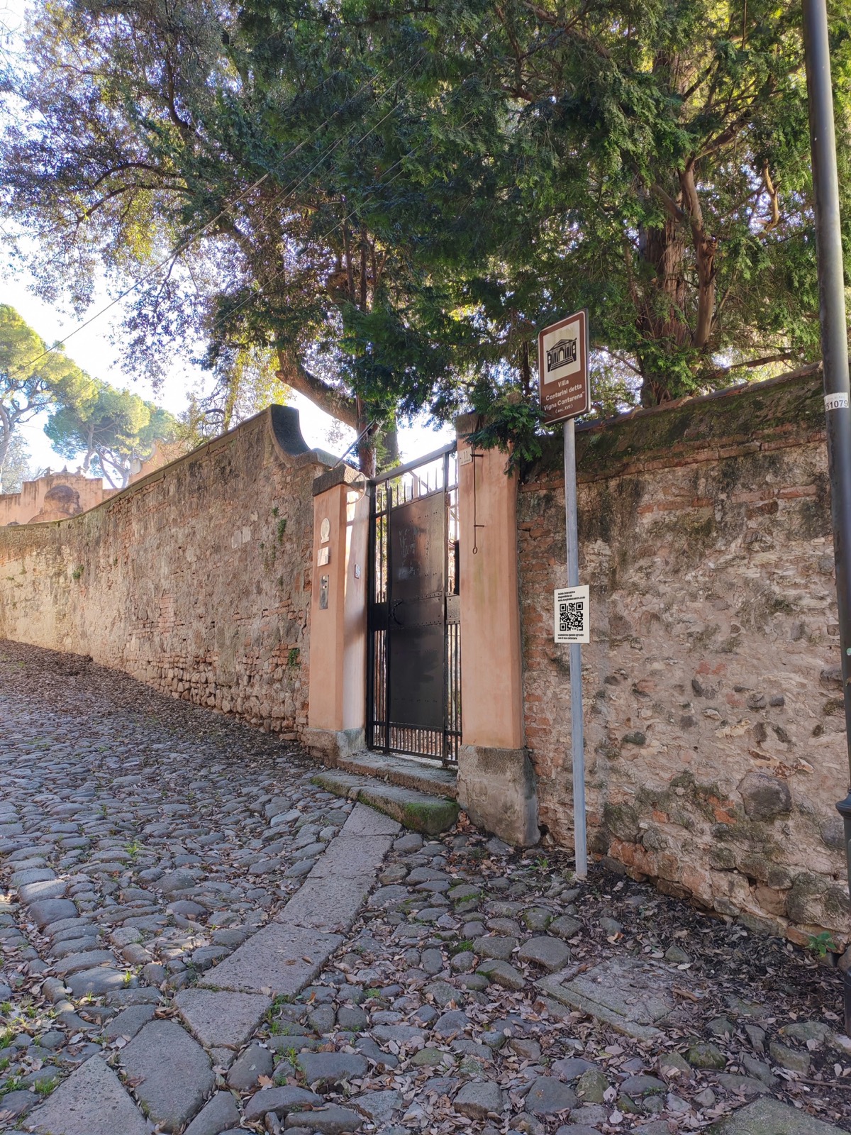 Simulazione: una targa con QR code su un palo lungo una calle acciottolata, accanto al cartello di una villa