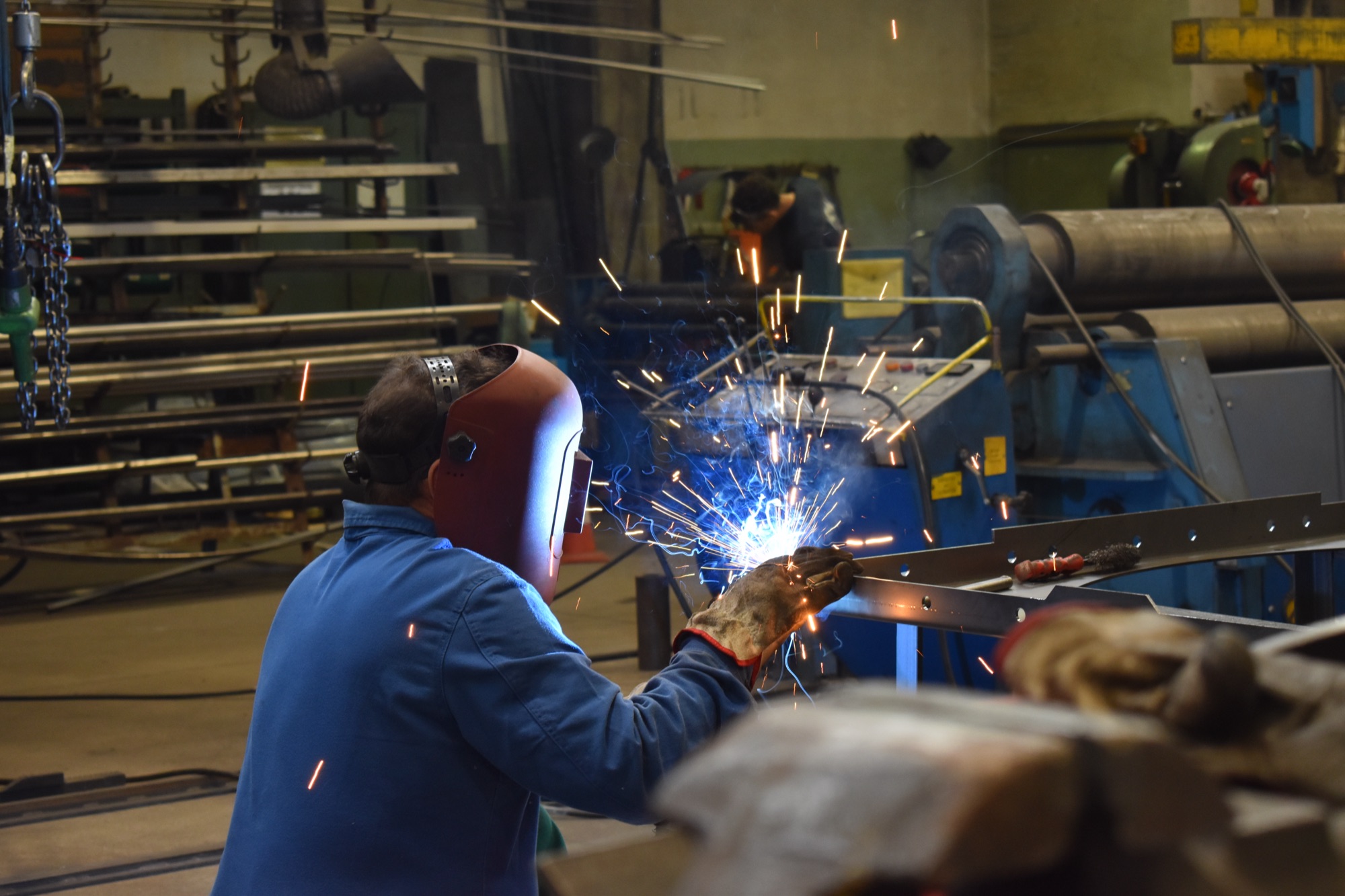 Un saldatore in tuta blu e casco rosso salda una trave in officina, fra la luce dell’arco e le scintille