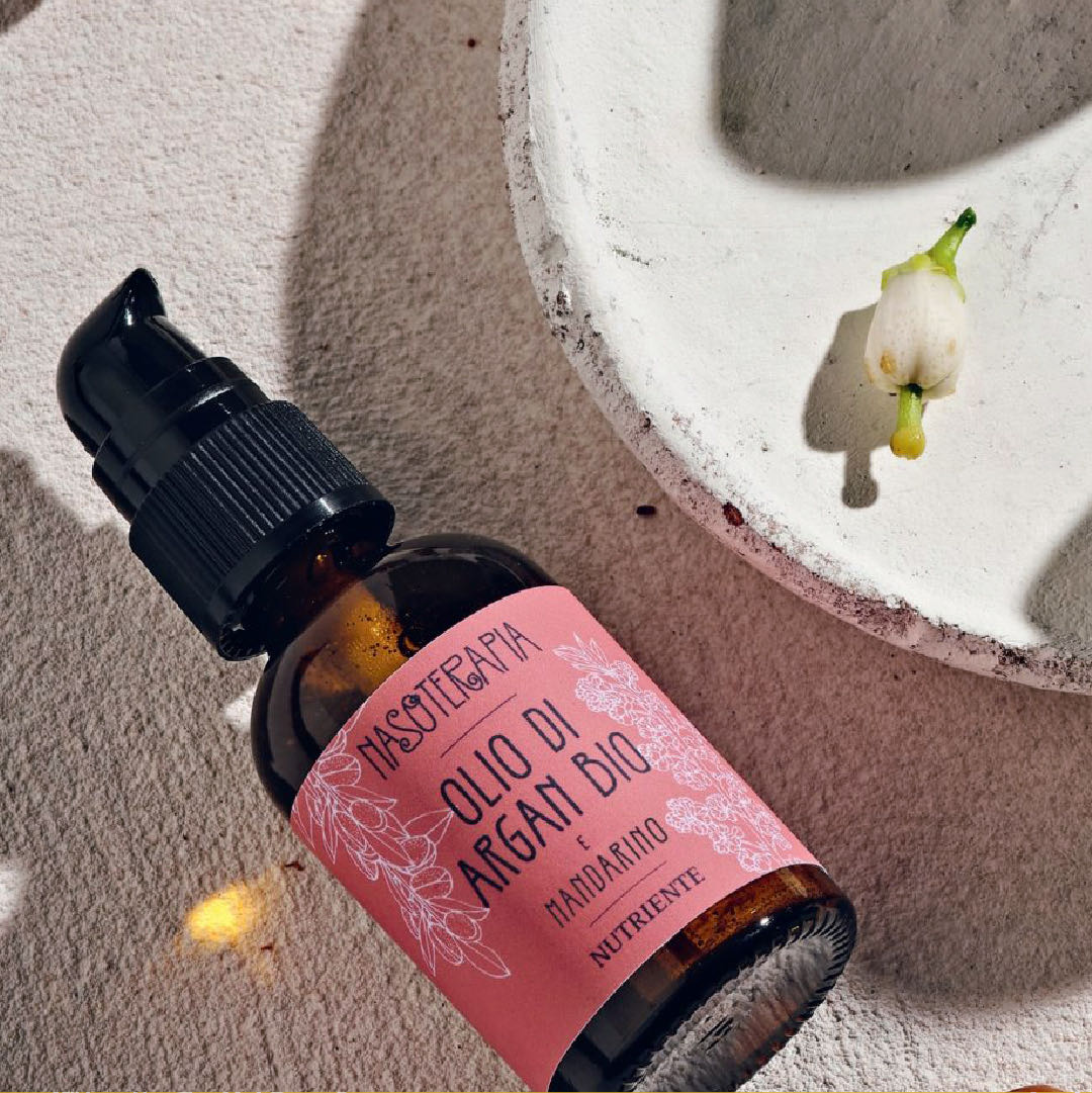 Dettaglio del flacone di olio di argan bio e mandarino, con l’etichetta rosa, accanto a un fiore