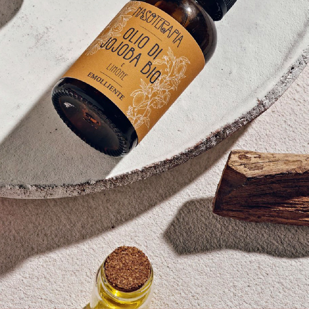 Dettaglio del flacone di olio di jojoba bio e limone, con l’etichetta ocra, accanto a un pezzo di legno e a una fiala d’olio