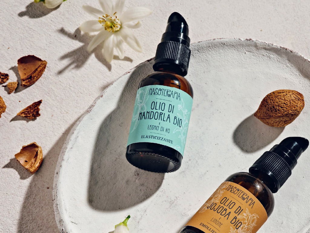 I flaconi di mandorla e jojoba con l’erogatore nero, su un piatto di ceramica bianca, accanto a una mandorla e a un fiore