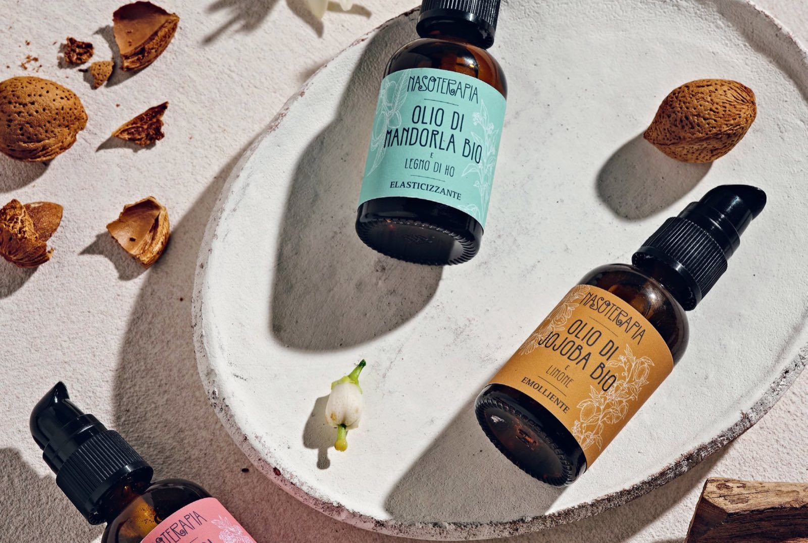I flaconi Nasoterapia di mandorla e jojoba con le etichette acqua e ocra, su un piatto di ceramica bianca tra mandorle e fiori