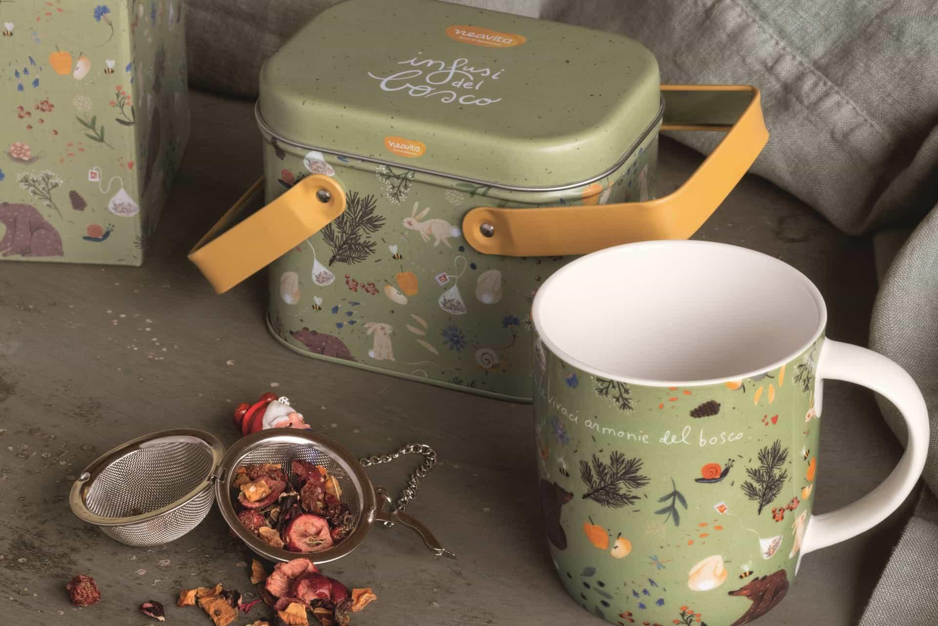 The green suitcase-style Infusi del bosco tin with its matching mug, the strainer and infusion petals on the table