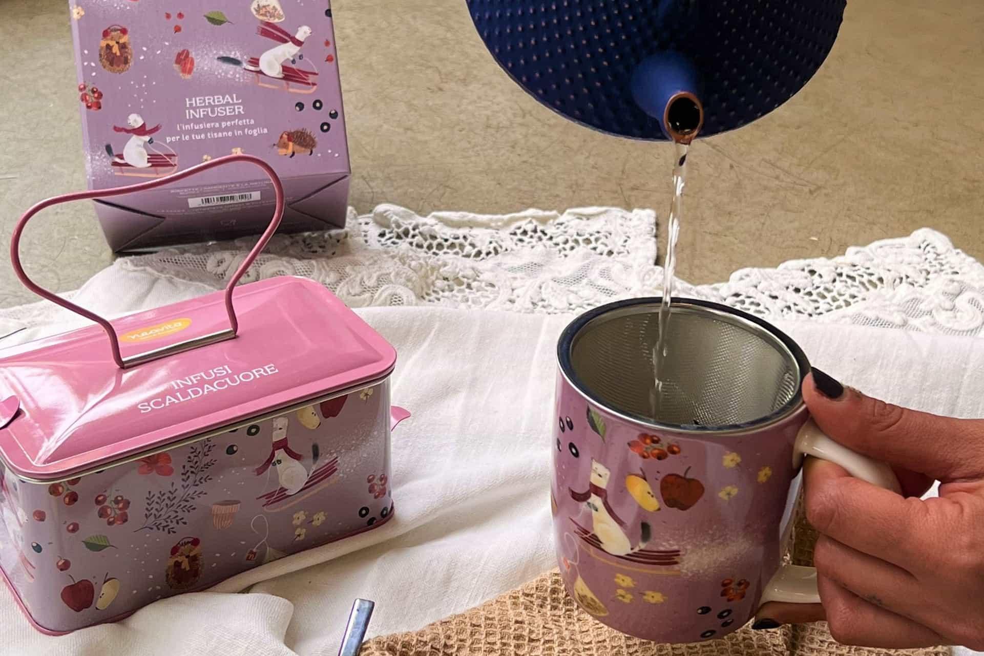 The Infusi scaldacuore line: pink tin with a handle, a mug with a strainer as water is poured in, and the infuser box