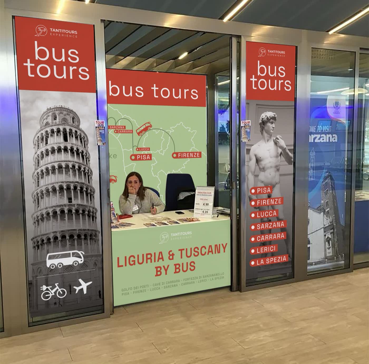 Simulazione del desk Tantitours 2022 nel terminal: laterali rossi con Torre di Pisa e David, vetro verde con la mappa, bancone Liguria & Tuscany by bus